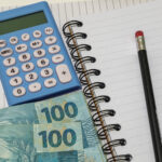 Uma calculadora ao lado de um lápis e duas notas de 100 reais